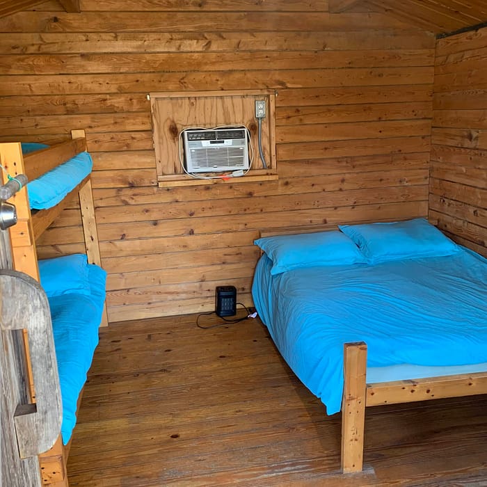A cozy wooden cabin room with a bunk bed, a double bed, blue bedding, and an air conditioning unit.