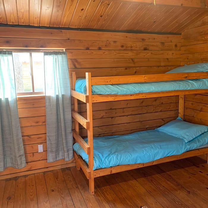 A wooden bunk bed with blue bedding in a cabin room with a window draped in light curtains.