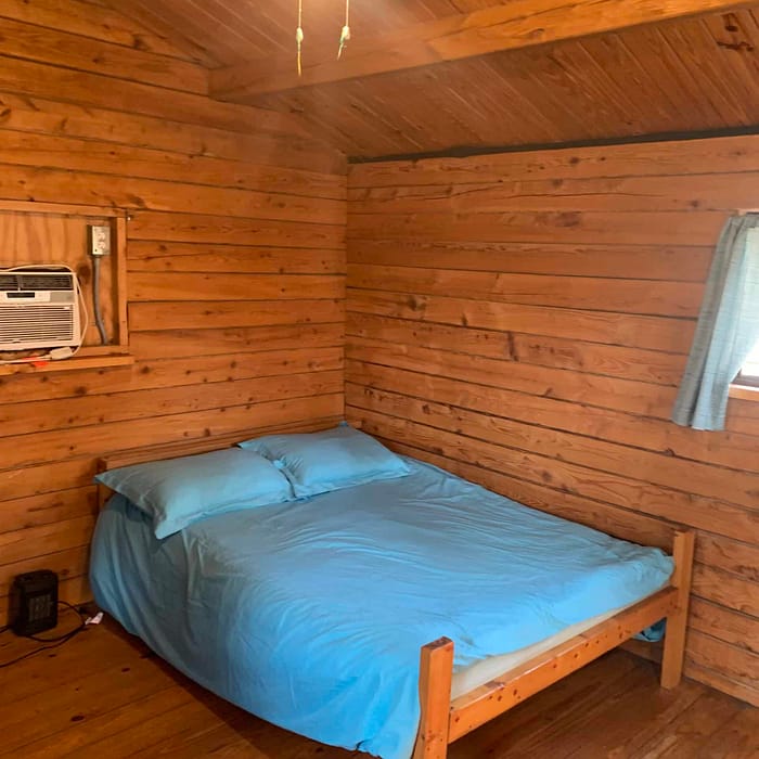 A small wooden cabin bedroom with a double bed, blue bedding, an air conditioner, and a ceiling fan.