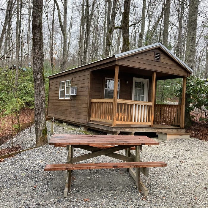 A wooden cabin with a front porch situated among trees, with a picnic table in the foreground.