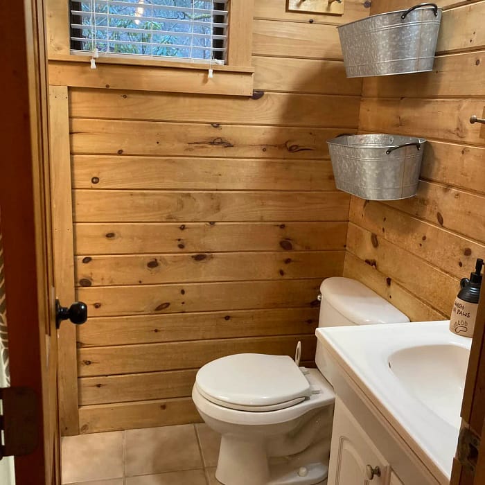 Rustic bathroom interior with wood-paneled walls, a white toilet beside a vanity sink, and galvanized buckets above as decor.