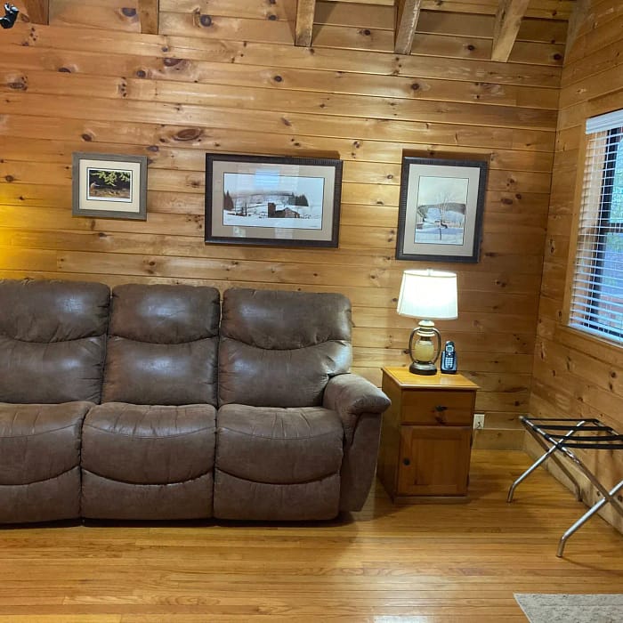 A cozy wooden cabin interior with a plush brown sofa, a lamp on a small table, and various pictures on the walls.
