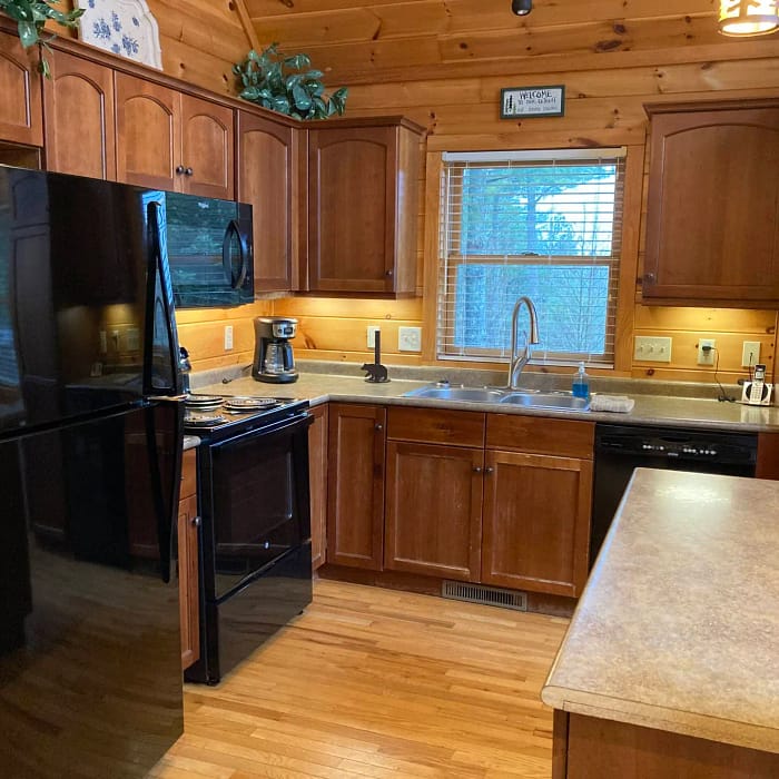 A cozy kitchen with wooden cabinets and floors features a black refrigerator, stove, and dishwasher. A coffee maker sits on the countertop near the sink, and a window above the sink allows natural light to brighten the space.