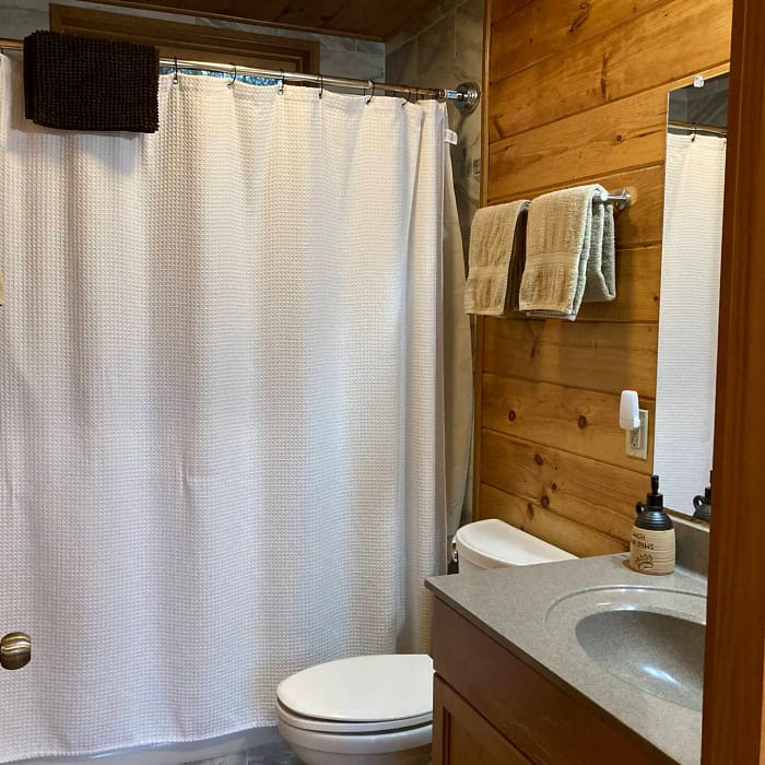 A small bathroom with wooden walls features a white shower curtain, a white toilet, and a wooden vanity with a grey countertop. Above the vanity is a mirror, and two beige towels are hanging on a rack to the right of the shower. A dark towel is draped over the curtain rod.