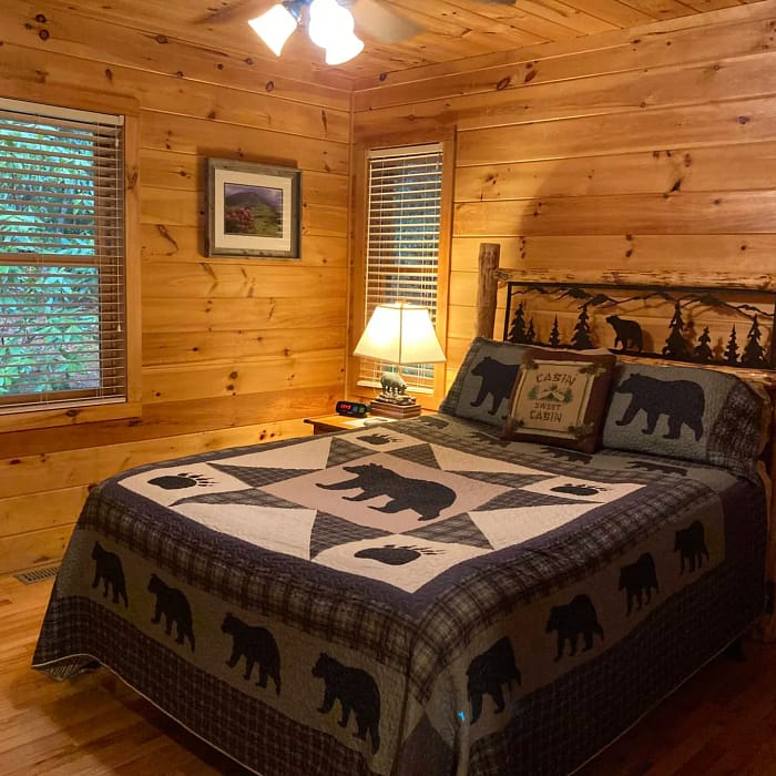 A cozy, rustic bedroom with wood paneling, bear-themed bedding, and forest view through the blinds.