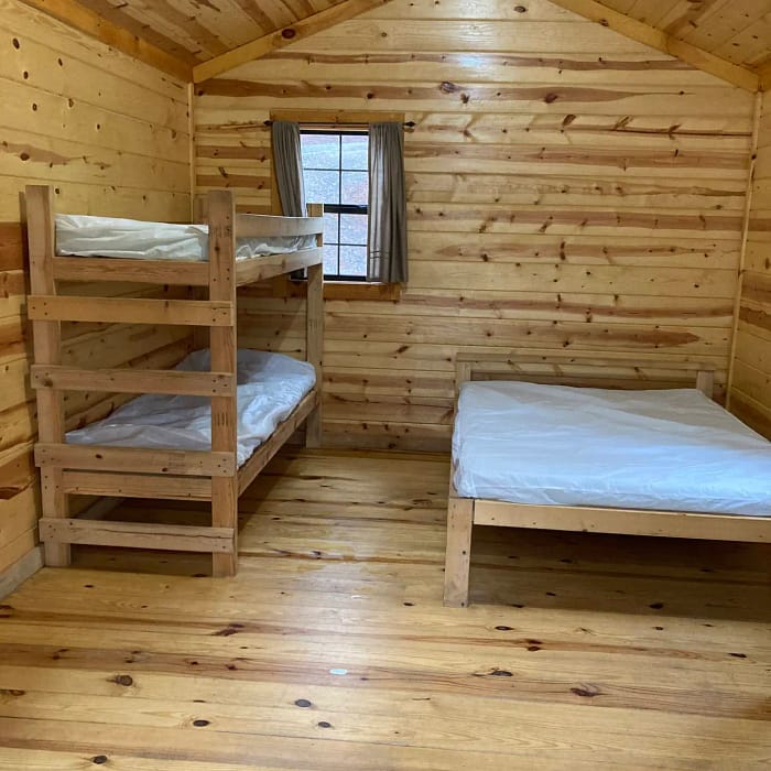 A small wooden cabin room with a bunk bed to the left and a single bed to the right, both with white mattresses.