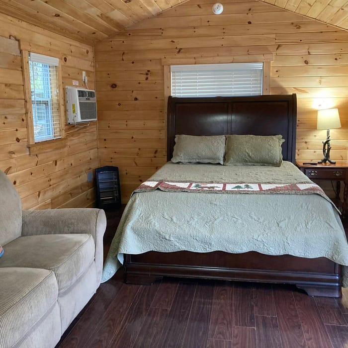 A cozy wooden cabin bedroom featuring a double bed with a quilt, side tables, an armchair, and an air conditioning unit.