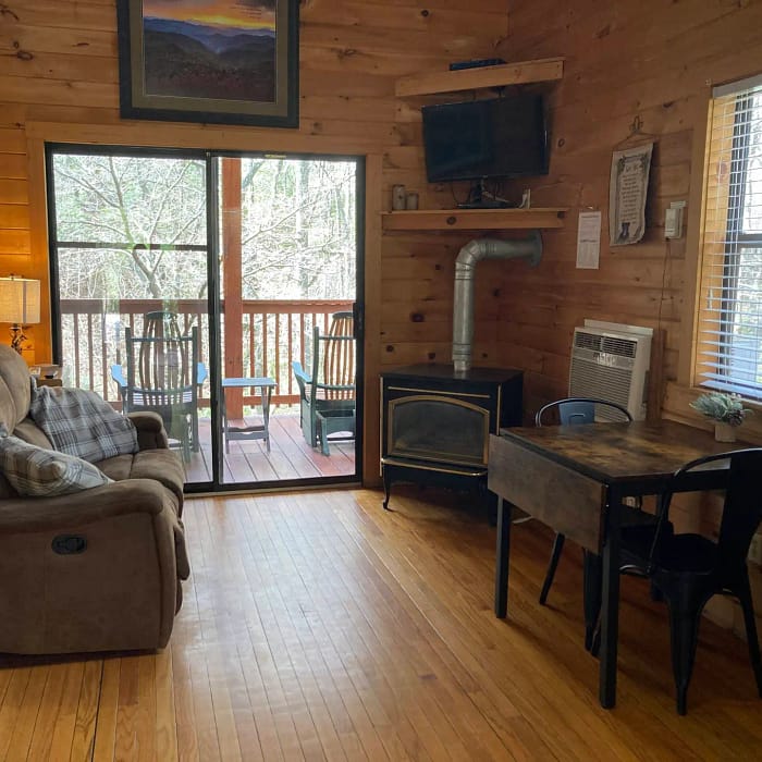 A cozy cabin interior with wooden walls, a recliner, a stove, a small dining table, and a deck with a view of the woods through large windows.