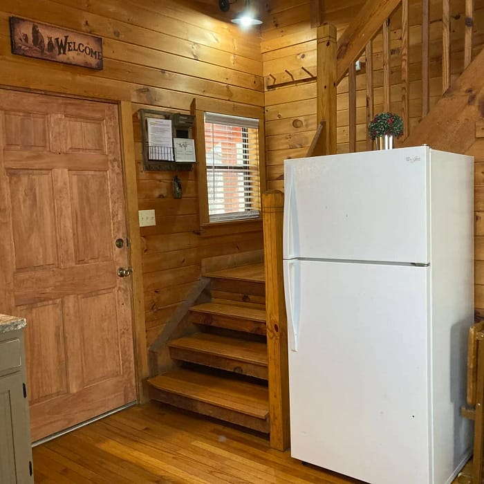 A cozy wooden cabin interior featuring a staircase, a refrigerator, and a door with a "welcome" sign above it.