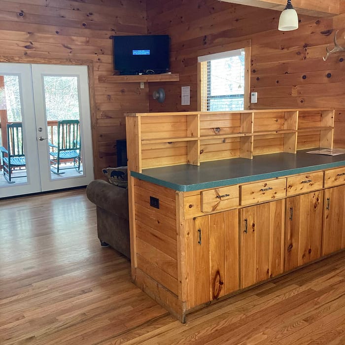 A cozy wooden cabin interior featuring a kitchen bar counter and a living area with a mounted tv and access to a balcony.