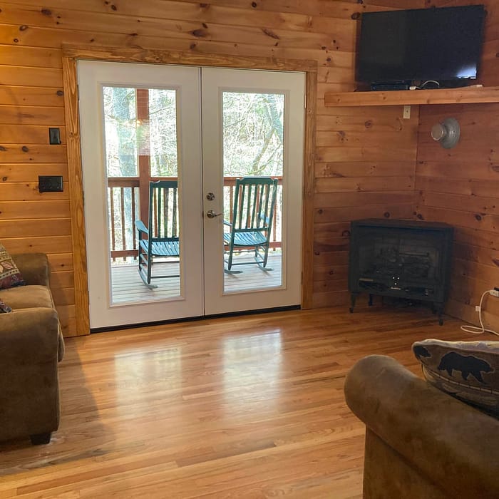 Cozy wooden cabin interior with a fireplace, comfortable seating, and glass doors leading to an outdoor deck.