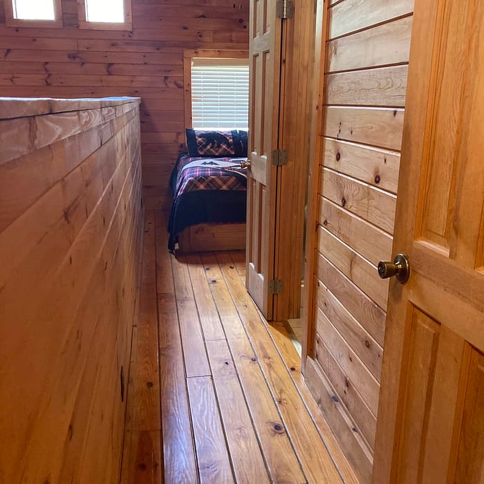 A wooden interior of a hallway leading to a bedroom, featuring hardwood floors, paneled walls, a ceiling fan, and an open door.