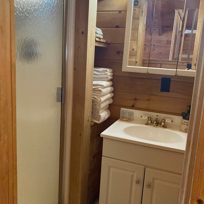 A bathroom interior with a wooden finish, featuring a sink vanity with a mirrored medicine cabinet and a stack of towels by a frosted glass shower door.