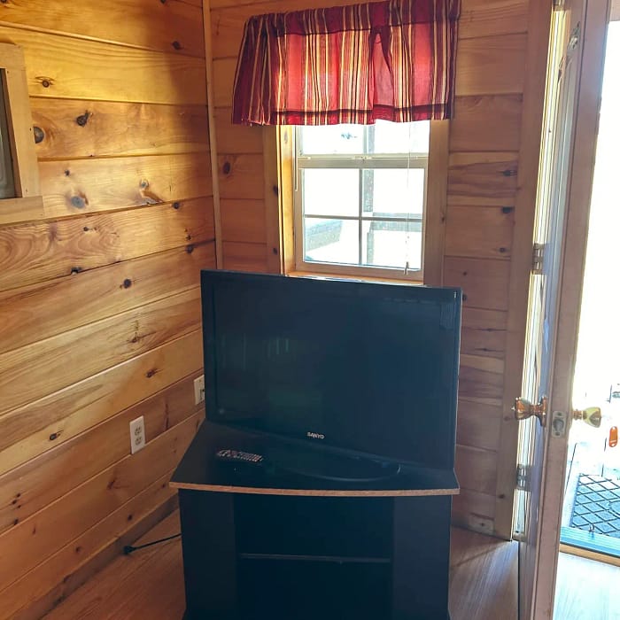 A small wooden room with a flat-screen TV on a black stand positioned in the corner, under a window with a red and yellow striped valance. Light streams in from a partially open door on the right, casting a warm glow on the wooden floor and walls.