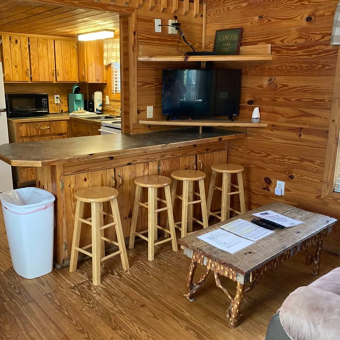 A cozy, wooden cabin interior with a kitchenette, bar stools, and a sitting area.