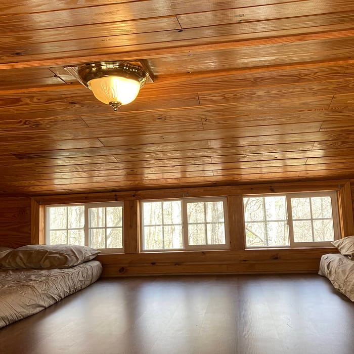 A cozy wooden cabin interior featuring a row of windows with a view of bare trees, two beds on either side, and a warm light fixture on the ceiling.