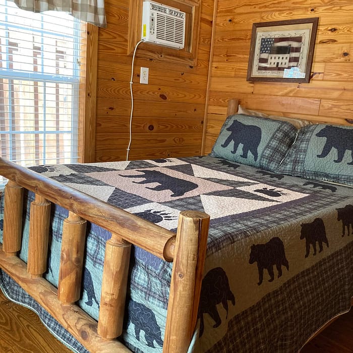 Wood-paneled bedroom with a rustic bed featuring bear motifs on the quilt and pillows, and an air conditioning unit above.