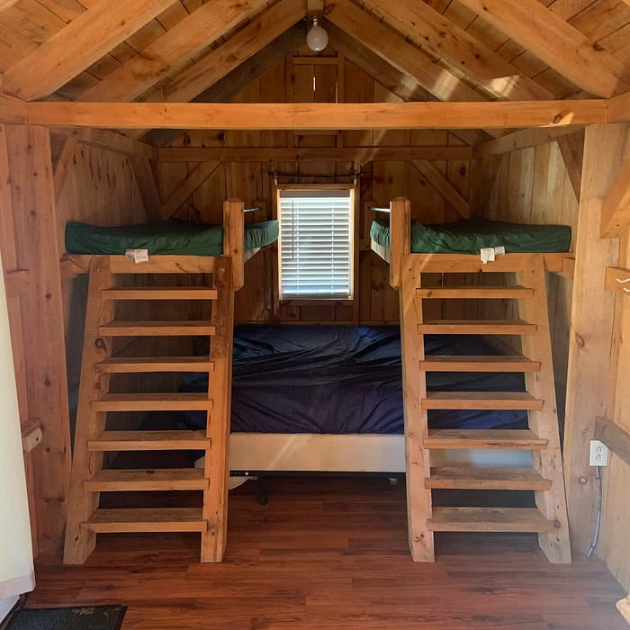 A cozy wooden cabin interior with two bunk beds flanking a window and a double bed below, featuring a ladder for upper bed access.