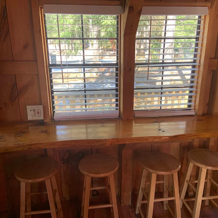 Wooden breakfast bar with three stools facing a window with horizontal blinds in a rustic cabin setting.