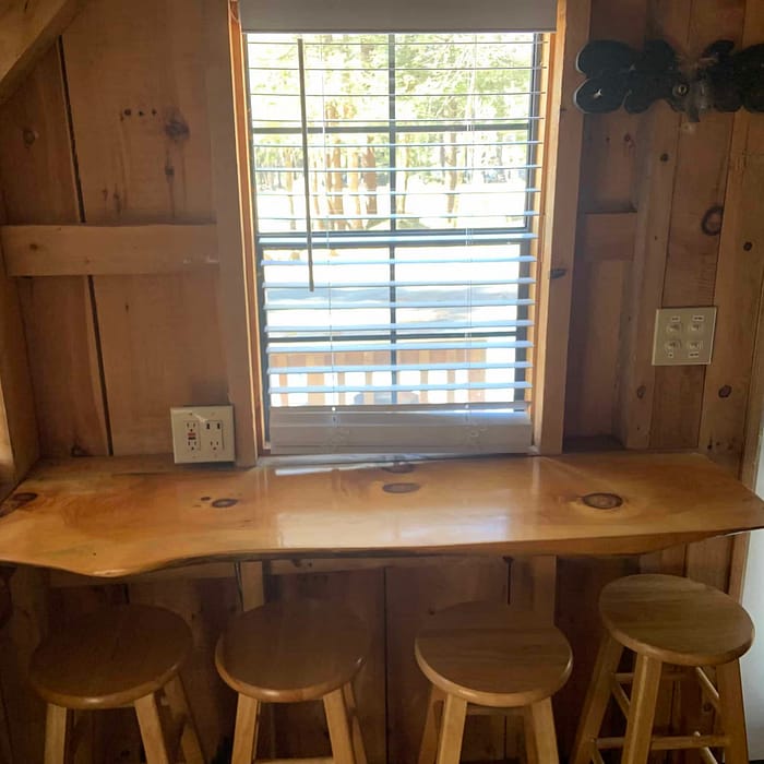 A wooden breakfast bar with stools by a window with blinds in a room with rustic wood paneling.