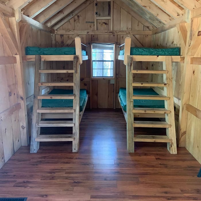 Interior of a wooden cabin with two bunk beds facing each other, a window at the far end, and a ceiling fan.