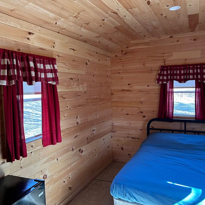 A small, cozy room with wooden paneled walls, a single bed with blue bedding, red and white curtains, and a mini-fridge.