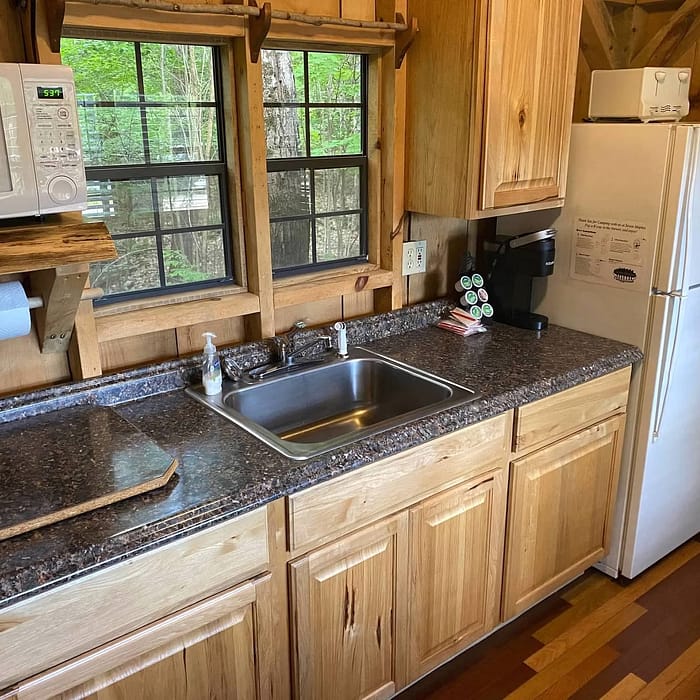 A compact kitchen interior with wooden cabinets, stainless steel sink, microwave, coffee maker, and a refrigerator next to a window with a view of trees.