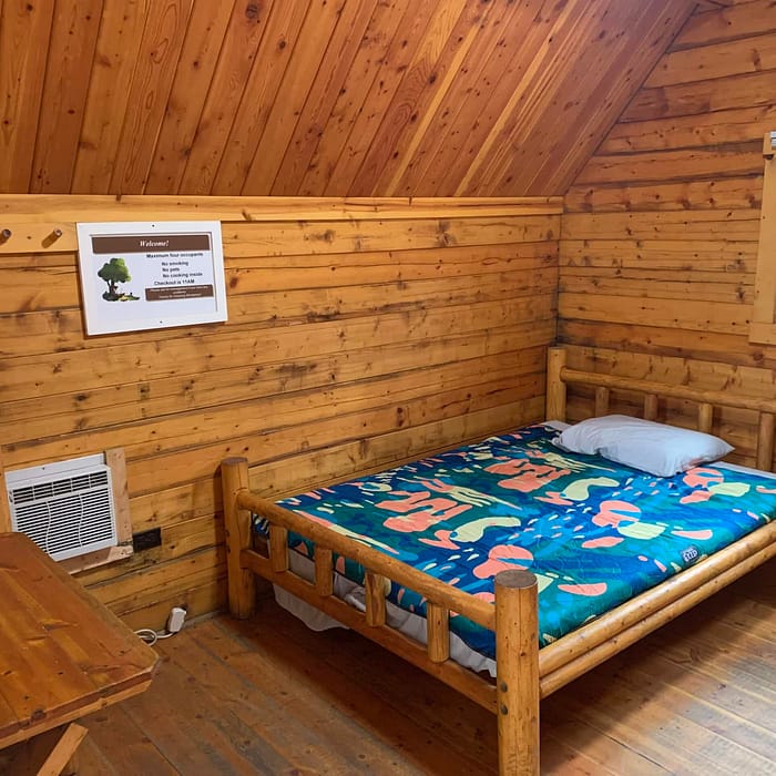 A simple wooden cabin interior with a single bed, colorful bedding, and an informational poster on the wall.
