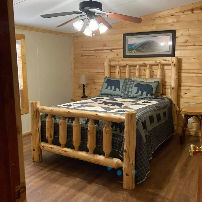 A rustic bedroom featuring a wooden bed with a bear-themed quilt, a ceiling fan, and wooden walls.