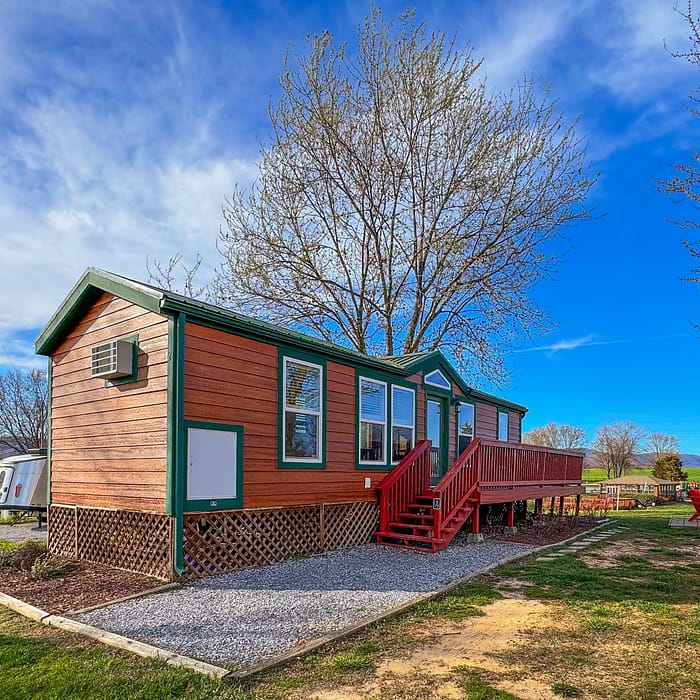 A small, single-story, green cabin with a red staircase and decking, located in an open outdoor area with clear skies above.