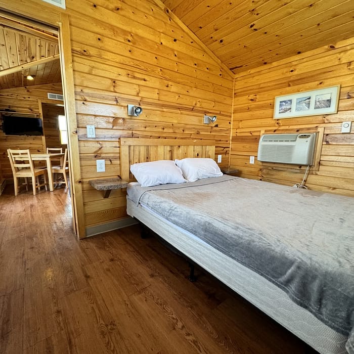 Rustic wooden cabin interior with a double bed, air conditioning, and a small dining area in the background.