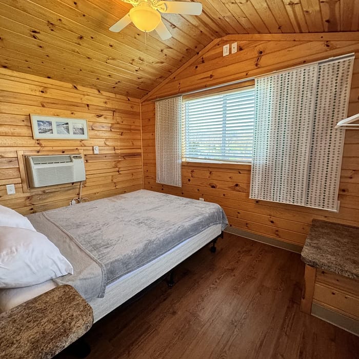 A cozy, rustic wooden cabin bedroom with a single bed, air conditioner, and ceiling fan.