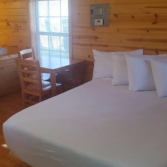 A cozy wooden cabin interior with a double bed dressed in white linens, a desk and chair near a window with blinds, and a bunk bed at the side. The walls are paneled with natural wood, creating a warm and rustic atmosphere.