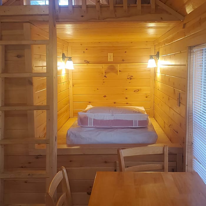 A cozy wooden cabin interior featuring a small lofted bed area with two wall-mounted reading lights. Below, there's a wooden dining table with two chairs. The walls and ceiling are entirely made of light-colored wood, creating a warm and inviting atmosphere.