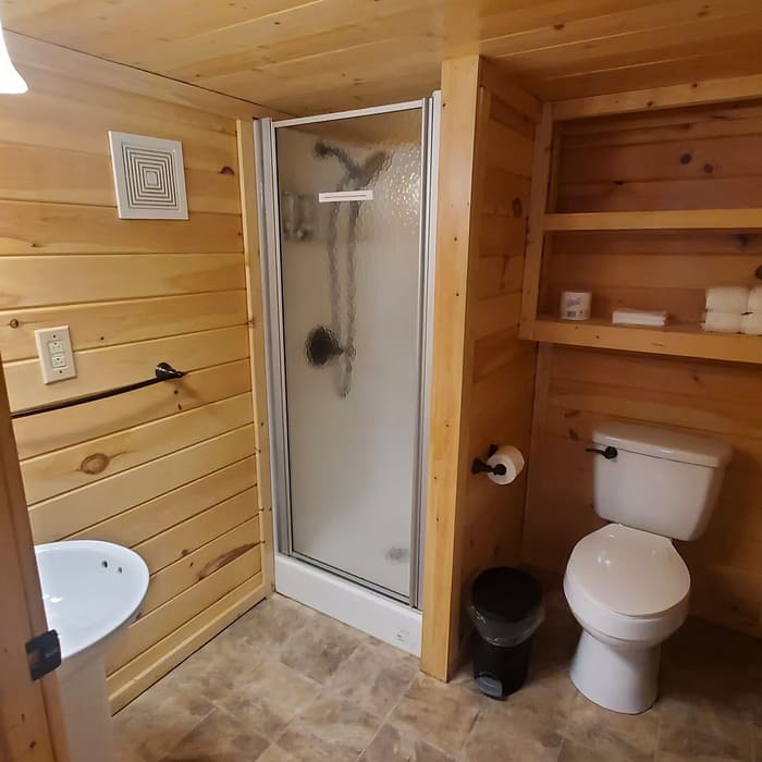 A small bathroom with wooden walls and ceiling, featuring a toilet, a corner shower with frosted glass doors, a sink with a mirror above it, and open shelves holding towels and toiletries. The floor is tiled with beige and brown tiles.