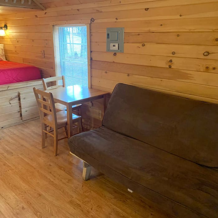 A cozy wooden cabin interior featuring a lofted twin bed with red bedding, a small dining table with two wooden chairs beneath a window with blinds, and a brown futon against the right wall on a wooden floor.