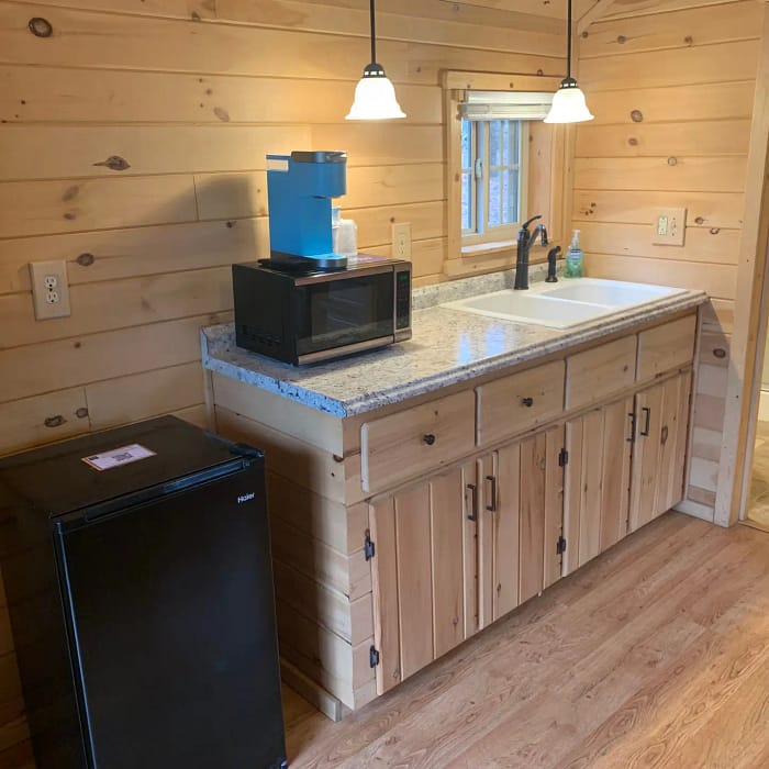 A quaint kitchenette with wooden walls features a granite countertop, a black microwave, and a blue coffee maker. A black mini-fridge stands beside the counter. Two pendant lights hang above the sink, and a window provides natural light. A door leads to an adjacent room.