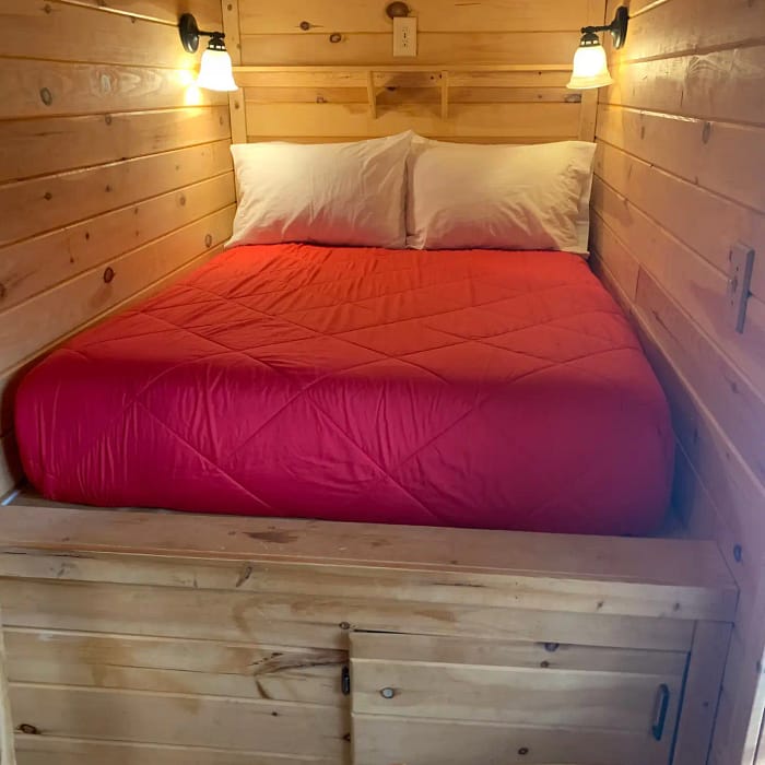 A cozy, wooden room with a double bed featuring a red quilt and two white pillows. The walls, floor, and ceiling are all made of light-colored wood. Two wall-mounted lamps are on either side of the headboard, illuminating the space warmly. A wooden chair is in the foreground.