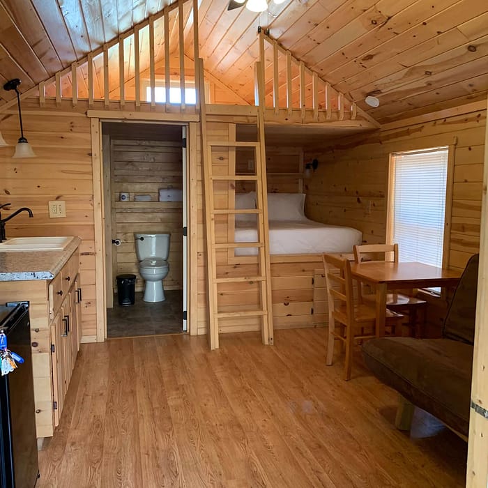 A small wooden cabin interior featuring a loft accessed by a ladder, a ceiling fan, and wooden walls and floors. The main area includes a kitchenette, a dining table with chairs, and a sofa. The back section has a bathroom with a white toilet and sink.