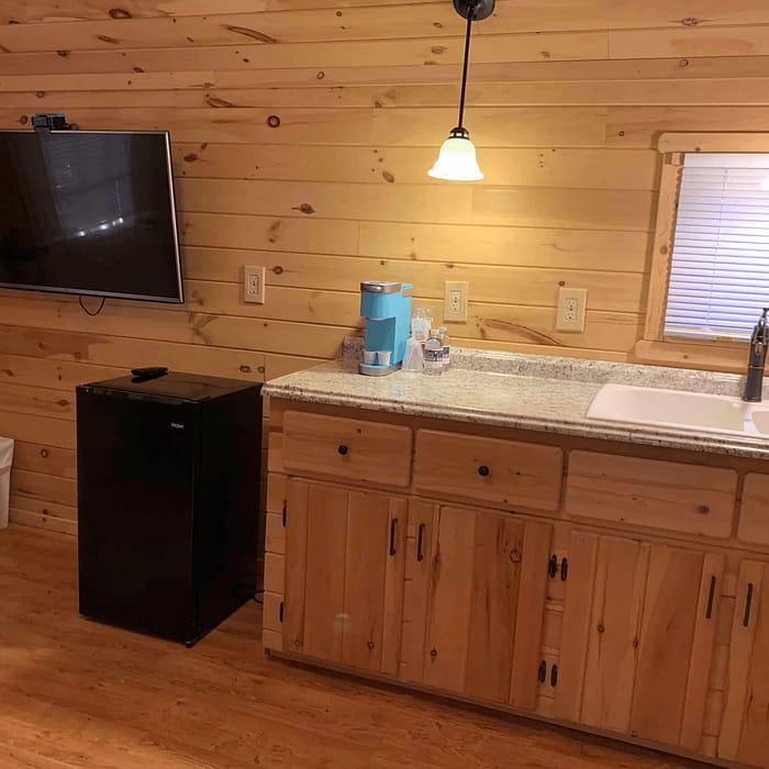A small room with wooden walls and flooring. A flat-screen TV mounted on the wall above a mini refrigerator. The countertop has a sink, a paper towel roll, and bottled water. Hanging lights illuminate the counter area. A small window with blinds is above the sink.