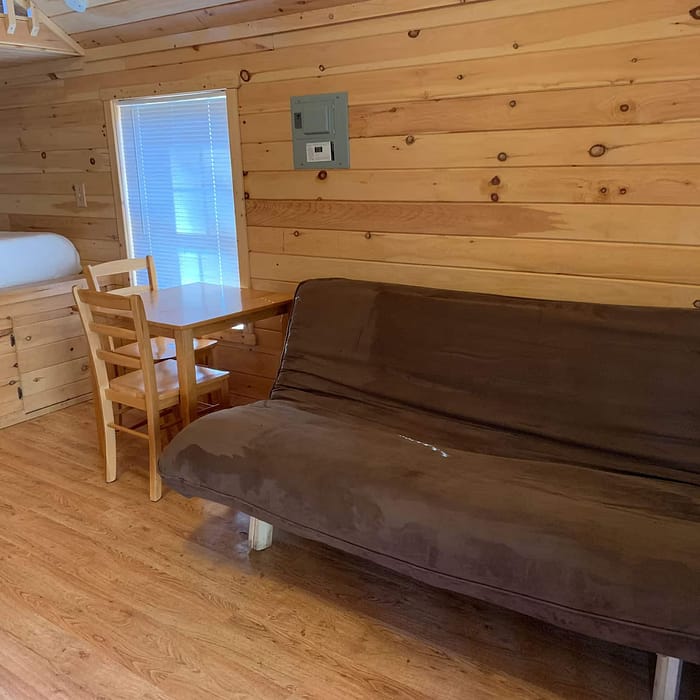 A cozy cabin interior with wooden walls and flooring. The room has a brown futon and a wooden desk paired with a matching chair. A bed with white linens occupies the corner, positioned above a wooden frame. A small window allows natural light in.