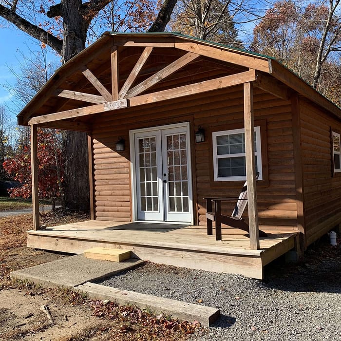 A small wooden cabin in a rural setting with a front porch, French doors, and a single window. The porch has a wooden bench and is surrounded by gravel and fallen leaves. Tall trees with sparse autumn foliage frame the background against a clear blue sky.