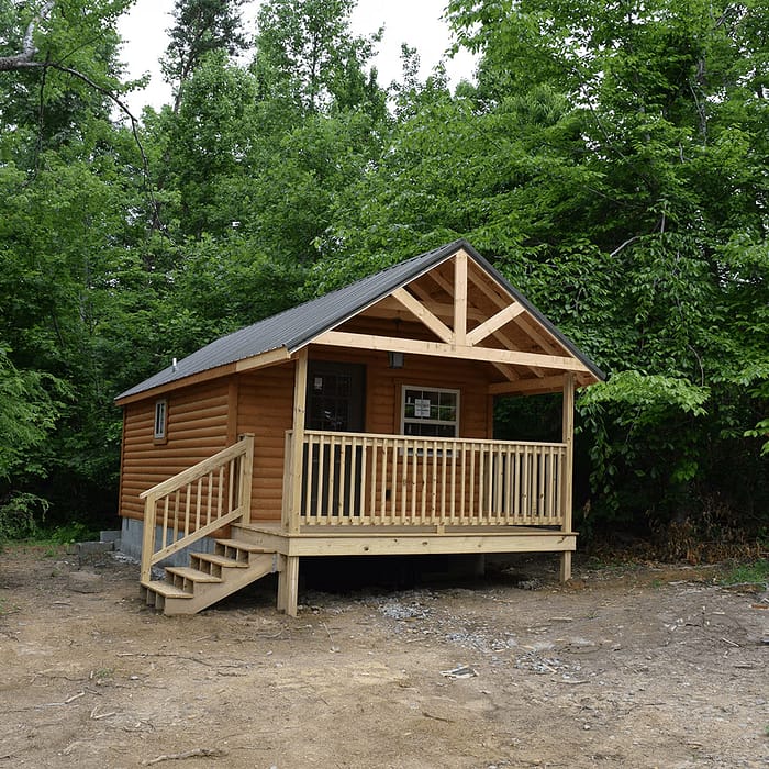 A small wooden cabin with a front porch and stairs is nestled among tall, leafy trees. The cabin, made of brown logs with a dark roof, is elevated on short stilts. The area around is clear from grass or gravel, highlighting the green forested background.