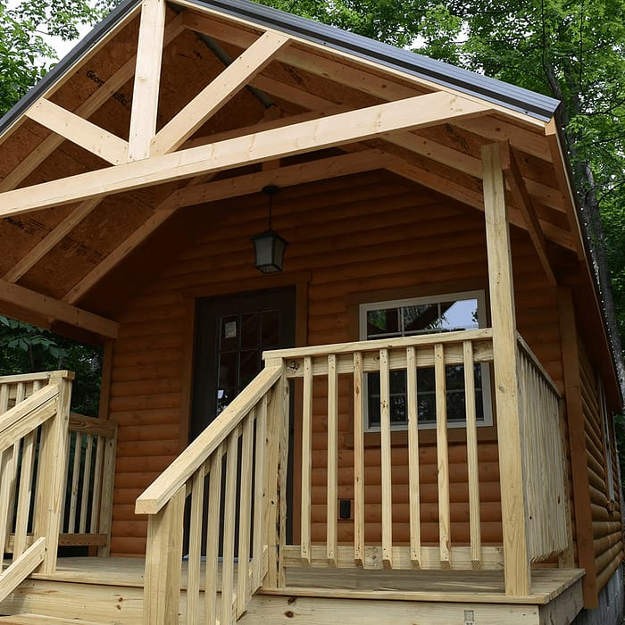 A small log cabin with a front porch stands amid lush green trees. The porch has wooden railings and steps leading up to a glass-paneled door. The cabin's roof has a triangular design with exposed beams, and a hanging light fixture is above the entrance.