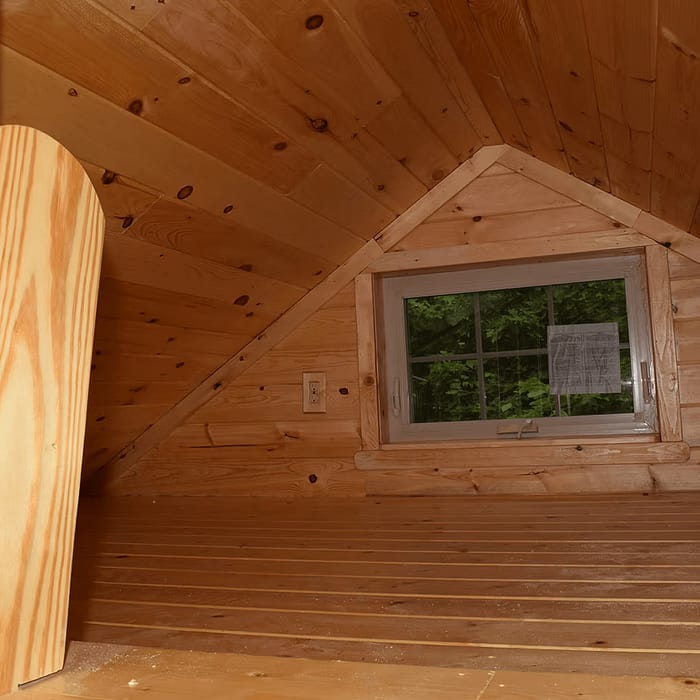 A small, cozy wooden attic with slanted ceilings, pine paneling, and a rectangular window offering a view of green foliage outside. The floor is wooden, and there is a power outlet on the wall near the window.