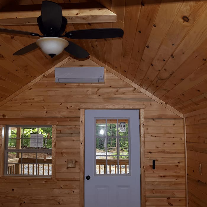 A cozy wooden interior features a ceiling fan with light, a small window to the left, and a white door with glass panes in the center. Soft-hued wood panels cover the walls and ceiling, with a conical pendant light hanging on the right side near the wall.