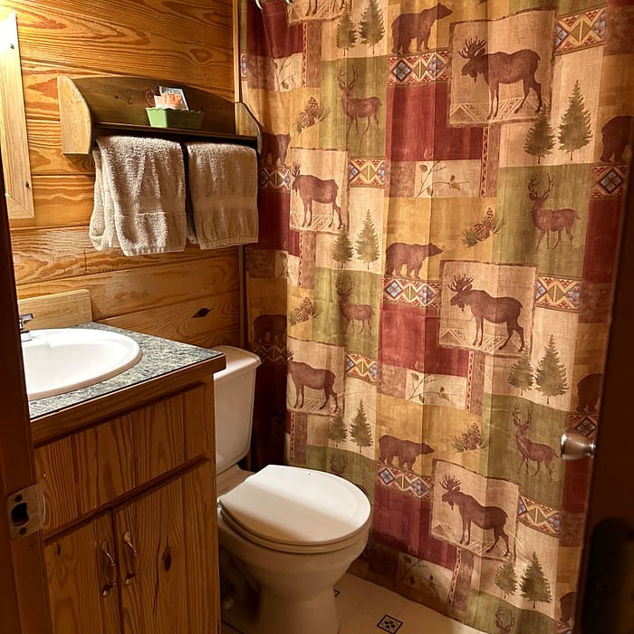 A cozy bathroom with wooden walls and ceiling, featuring a sink, toilet, and a shower with a moose-themed curtain in earthy tones. Above the toilet, a shelf holds folded towels and toiletries. The floor has a patterned, linoleum finish. Warm lighting is provided by a wall-mounted lamp.