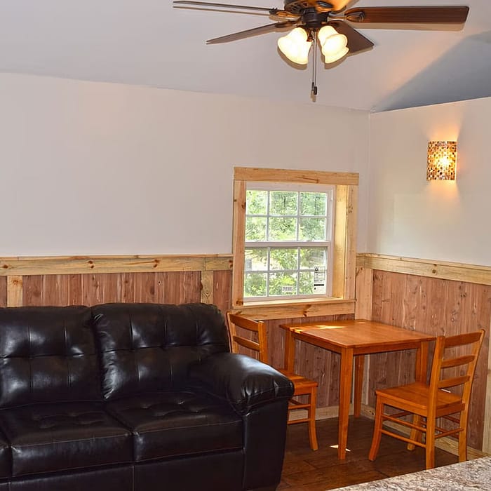 A cozy living space with a black leather couch, a wooden table with two chairs near a window, and a ceiling fan with lights. The room features wooden wainscoting on the lower half of the walls and a sconce light fixture next to the window.