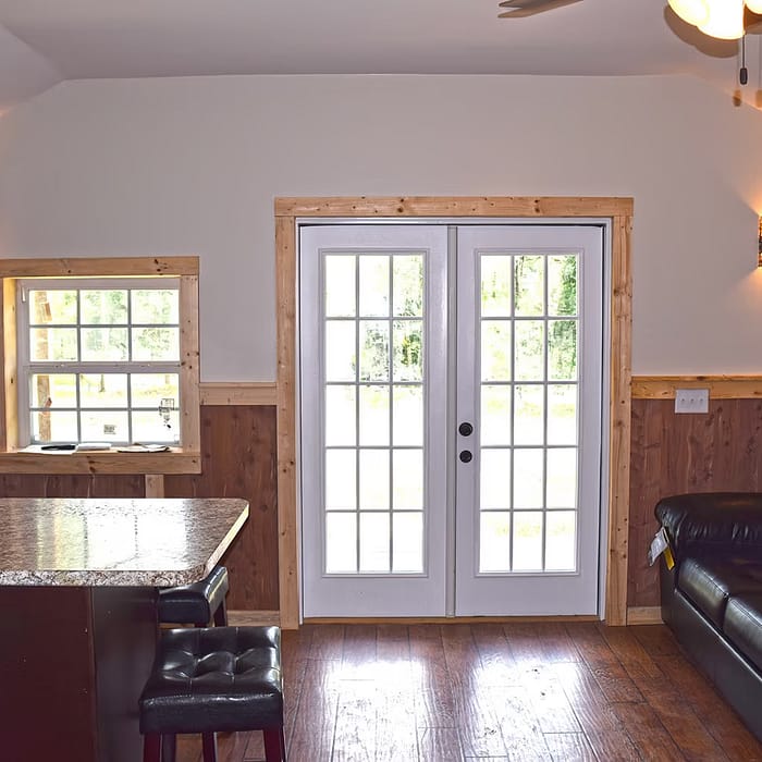 A cozy living room with a dark leather couch, a wall-mounted TV, a small window to the left, and French doors leading outside. There's a kitchen island with a granite countertop and two cushioned stools. Warm lighting and wooden accents create a rustic feel.