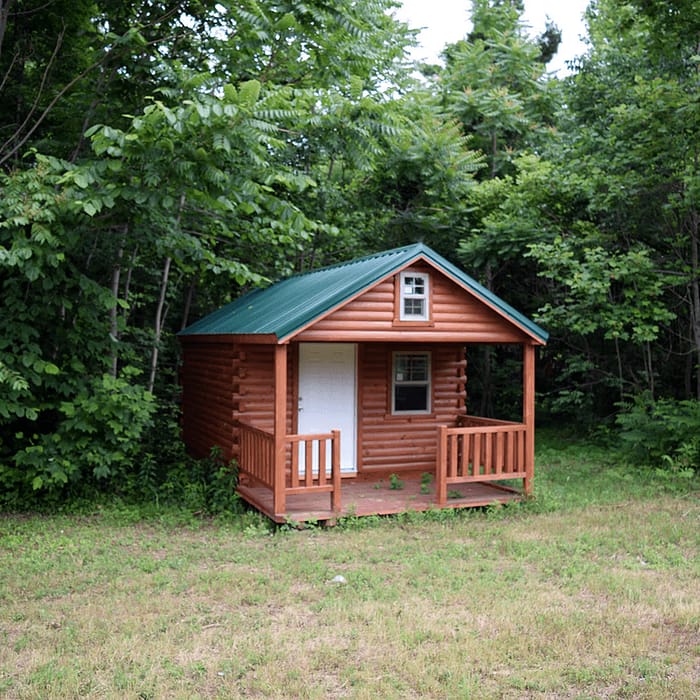 A small, rustic cabin with a green metal roof and wooden siding is situated in a forested area. The cabin has a tiny front porch and is surrounded by lush greenery and trees. The ground in front of the cabin is partially grass-covered.